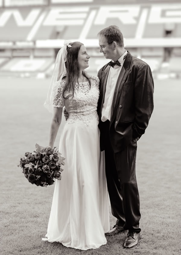 Ein Brautpaar steht auf einem Rasenplatz, die Braut trägt ein langes Kleid und hält einen Blumenstrauß, der Bräutigam trägt einen Anzug.