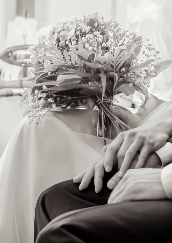 Ein Paar sitzt nebeneinander, die Hände ineinander verschlungen, mit einem Blumenstrauß im Hintergrund.