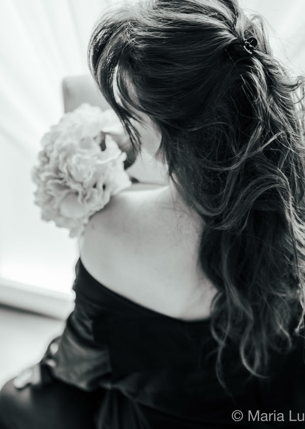 Frau mit langen Haaren, die einen Blumenstrauß hält, sitzt in einem schwarzen Kleid vor einem Fenster.