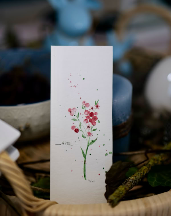 Ein rechteckiges Papier mit einer Zeichnung von rosa Blumen und grünen Blättern, umgeben von unscharfen Objekten und einem Korb im Hintergrund.