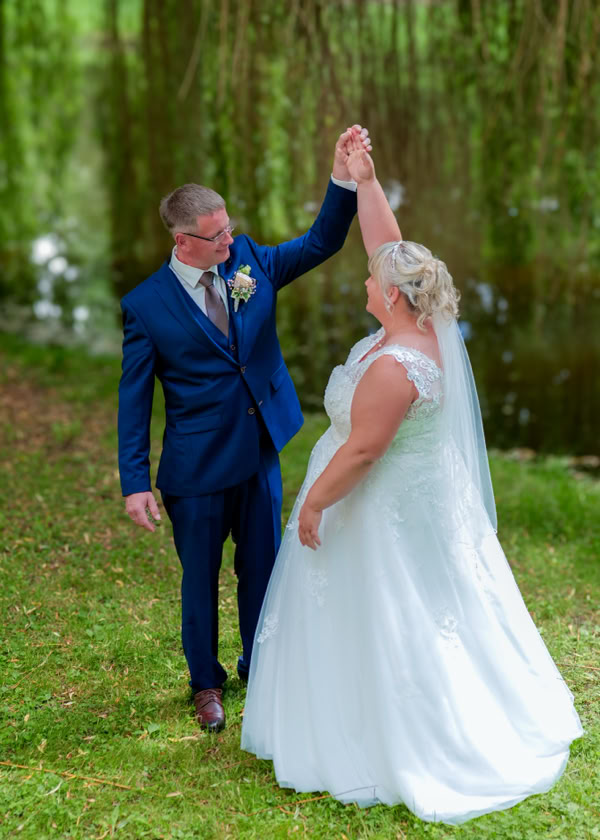 Ein Paar in Hochzeitskleidung tanzt auf einer Wiese vor einem Teich, umgeben von Bäumen.