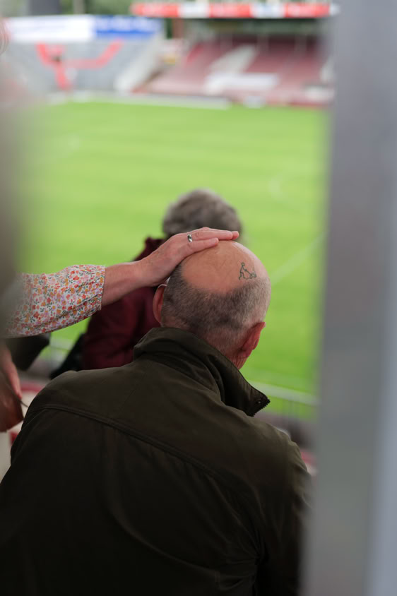 Eine Person legt die Hand auf den Kopf eines Mannes mit Glatze, im Hintergrund ist ein unscharfes Fußballfeld zu sehen.