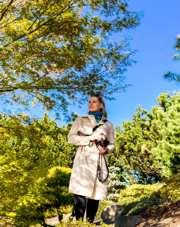 Eine Person in einem beigen Mantel steht in einem Garten mit Bäumen und Sträuchern unter einem klaren blauen Himmel.