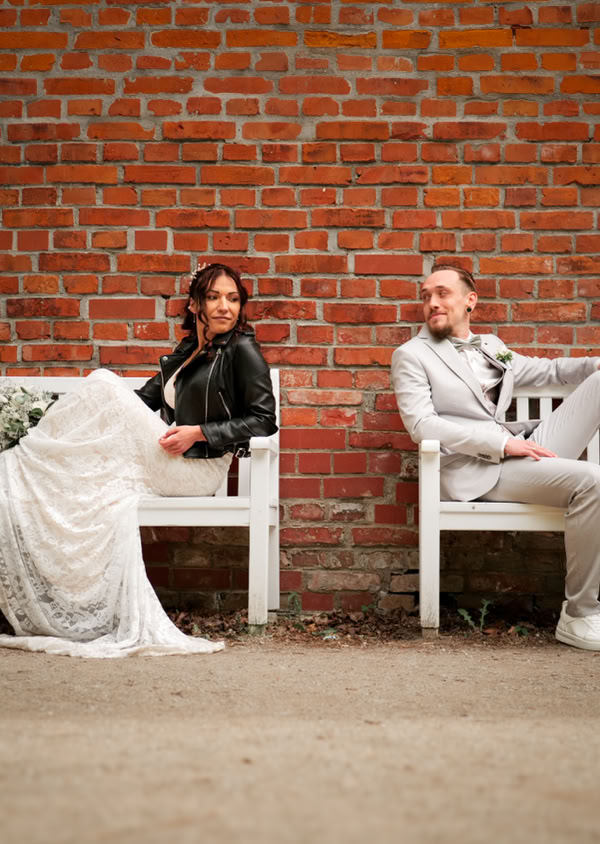 Zwei Personen sitzen auf weißen Bänken vor einer Ziegelmauer; eine Person trägt ein weißes Kleid und eine schwarze Jacke, die andere einen hellen Anzug.