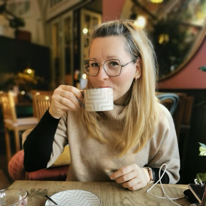 Eine Person in einem blauen Outfit steht auf einem Kiesweg neben einem Blumenbeet mit grünen Büschen und blauen Blumen, im Hintergrund eine Steintreppe.
